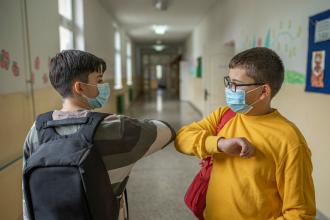 
		Zwei Schulkinder begrüßen sich in der Schule mit Masken und Elbogen.
	